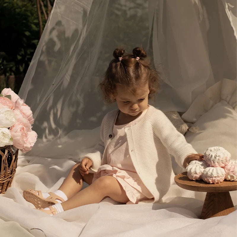 A young girl wearing pink Gladiator jelly sandals is sitting in a white fabric tent surrounded by soft light.