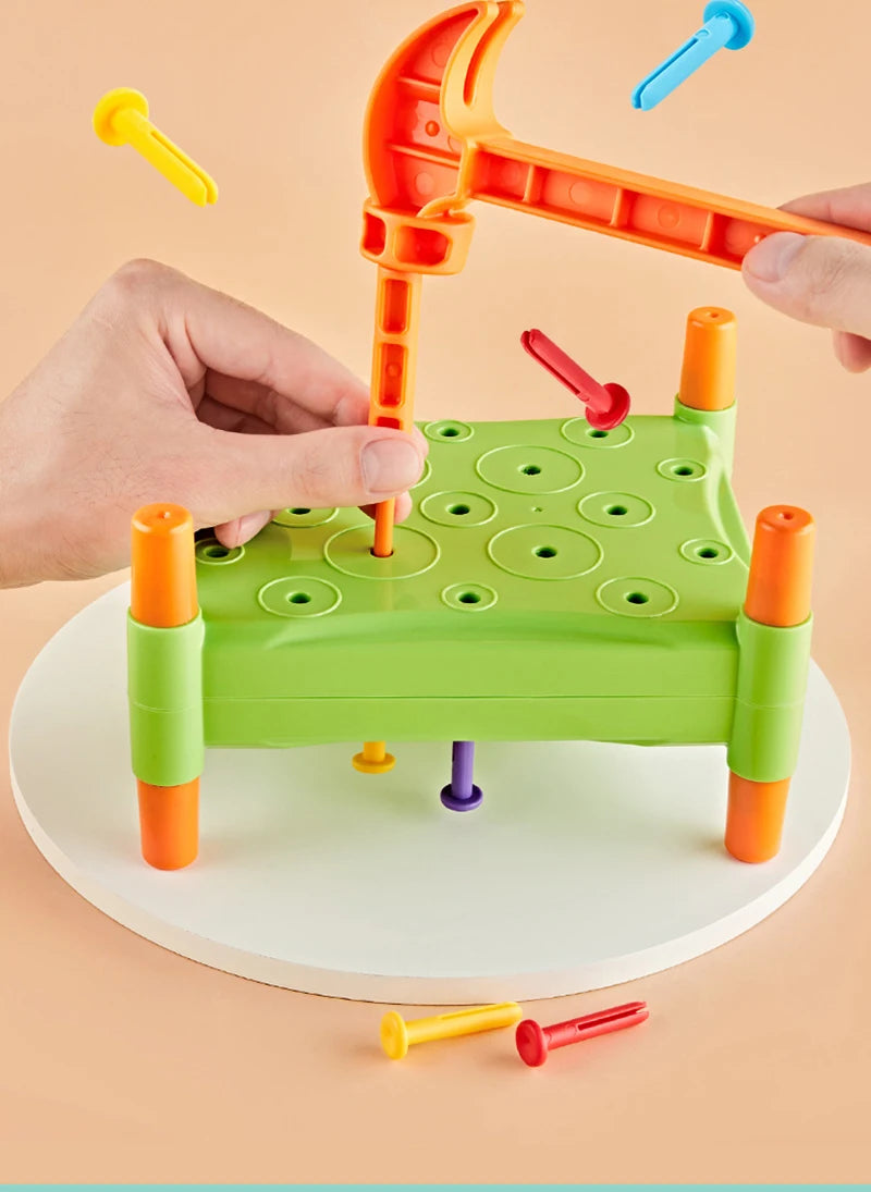 A hand is using an orange toddler hammer nails toy to insert colored wooden pegs into a green board.