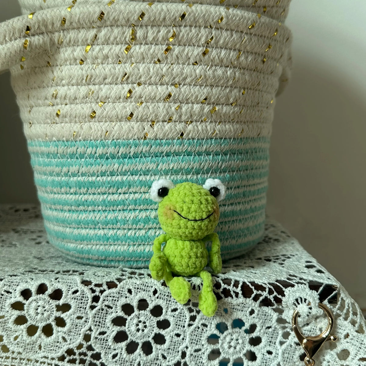 A handmade crochet frog keychain with green body and white eyes is displayed against a striped knitted bag charm.
