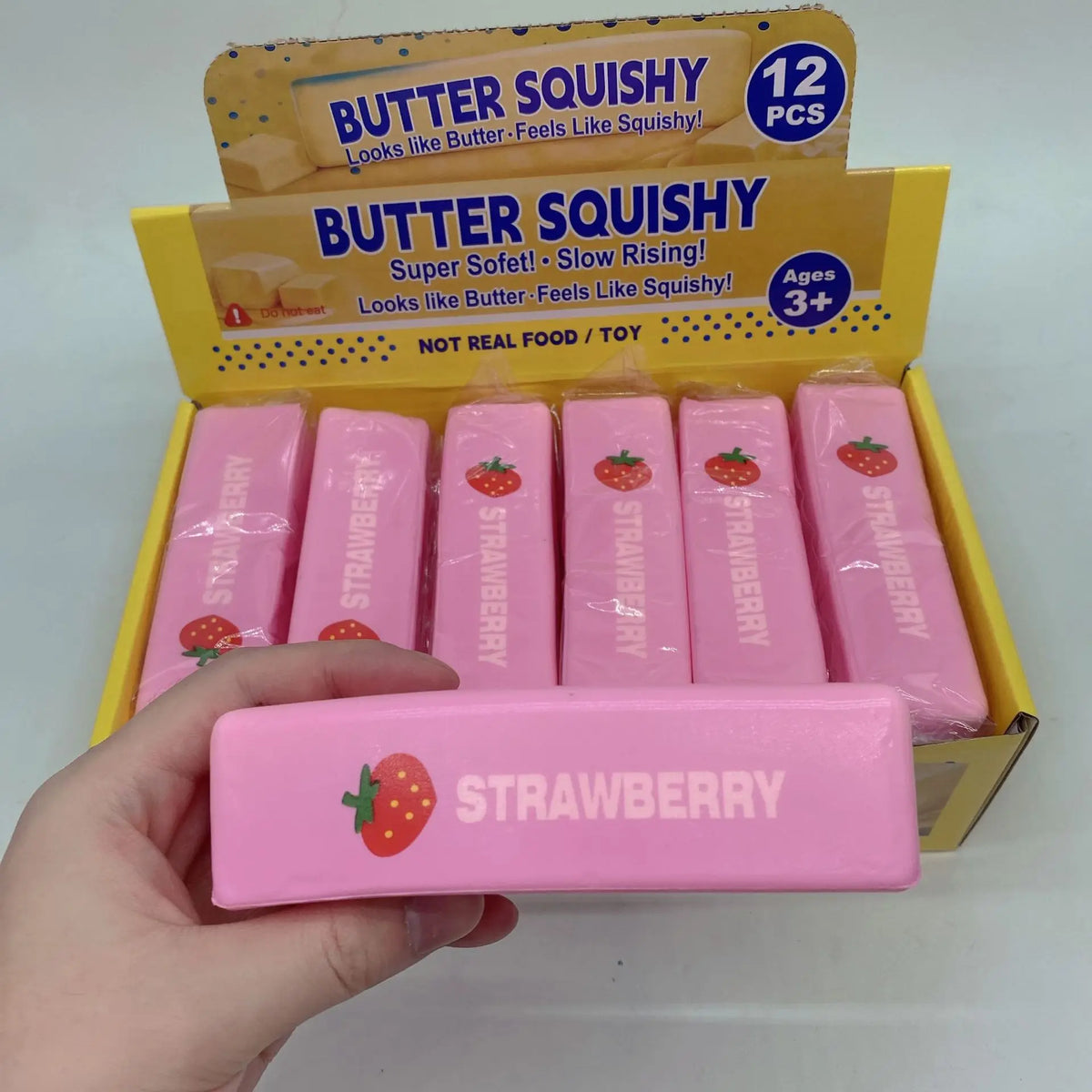 A hand holds a pink squishy strawberry stress relief toy packaged in a yellow box.