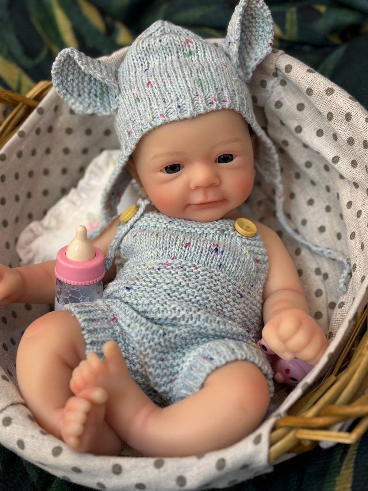 A hand-painted silicone reborn doll wearing a knitted elf hat and outfit, resting in a woven basket.
