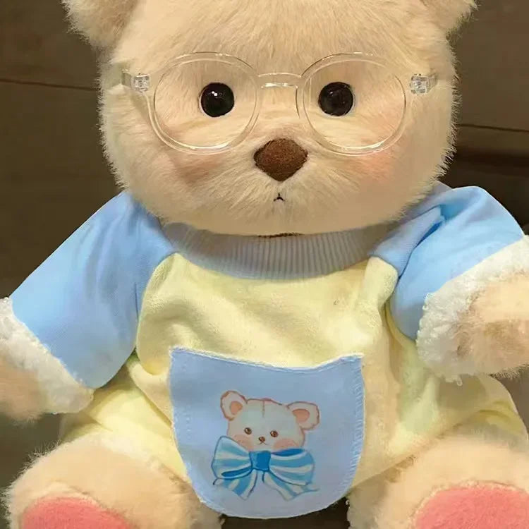 A fluffy teddy bear plush wearing a light blue and yellow dress with clear glasses.