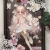 A detailed figure of a pink-haired girl in a frilly dress with white accents, displayed in a wooden frame surrounded by lace 
