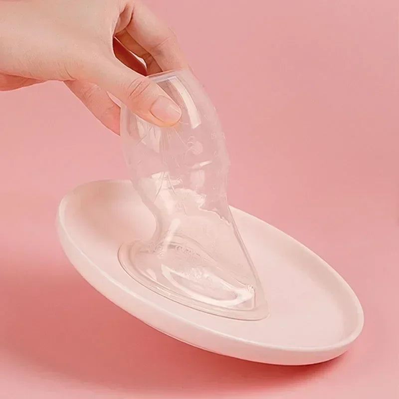 A hand is holding a clear manual breast pump for milk expression over a pink silicone plate.