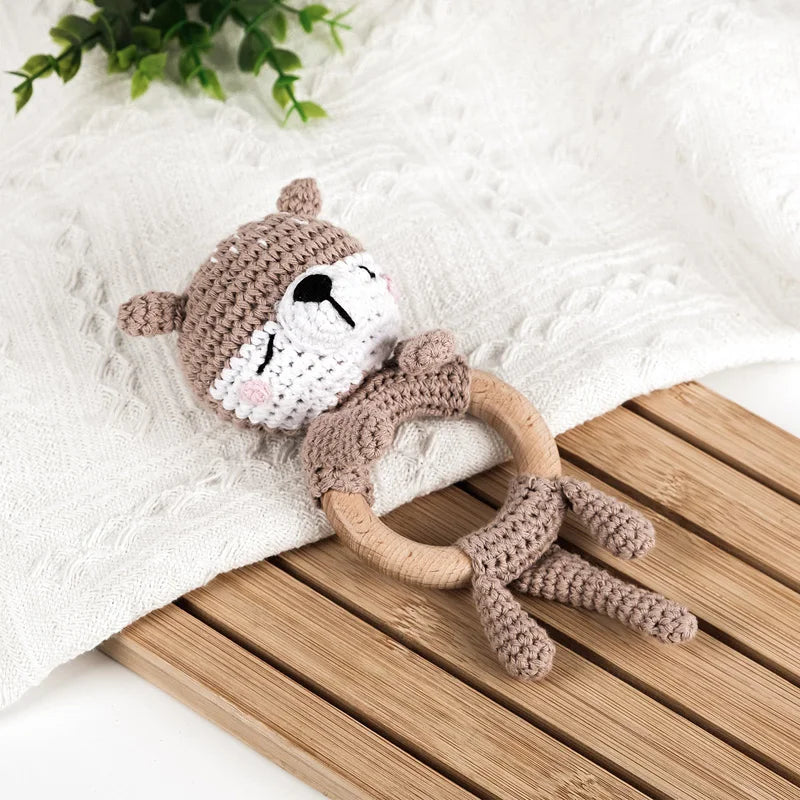 A brown knitted otter baby rattle and teether ring resting on a white blanket and wooden surface.
