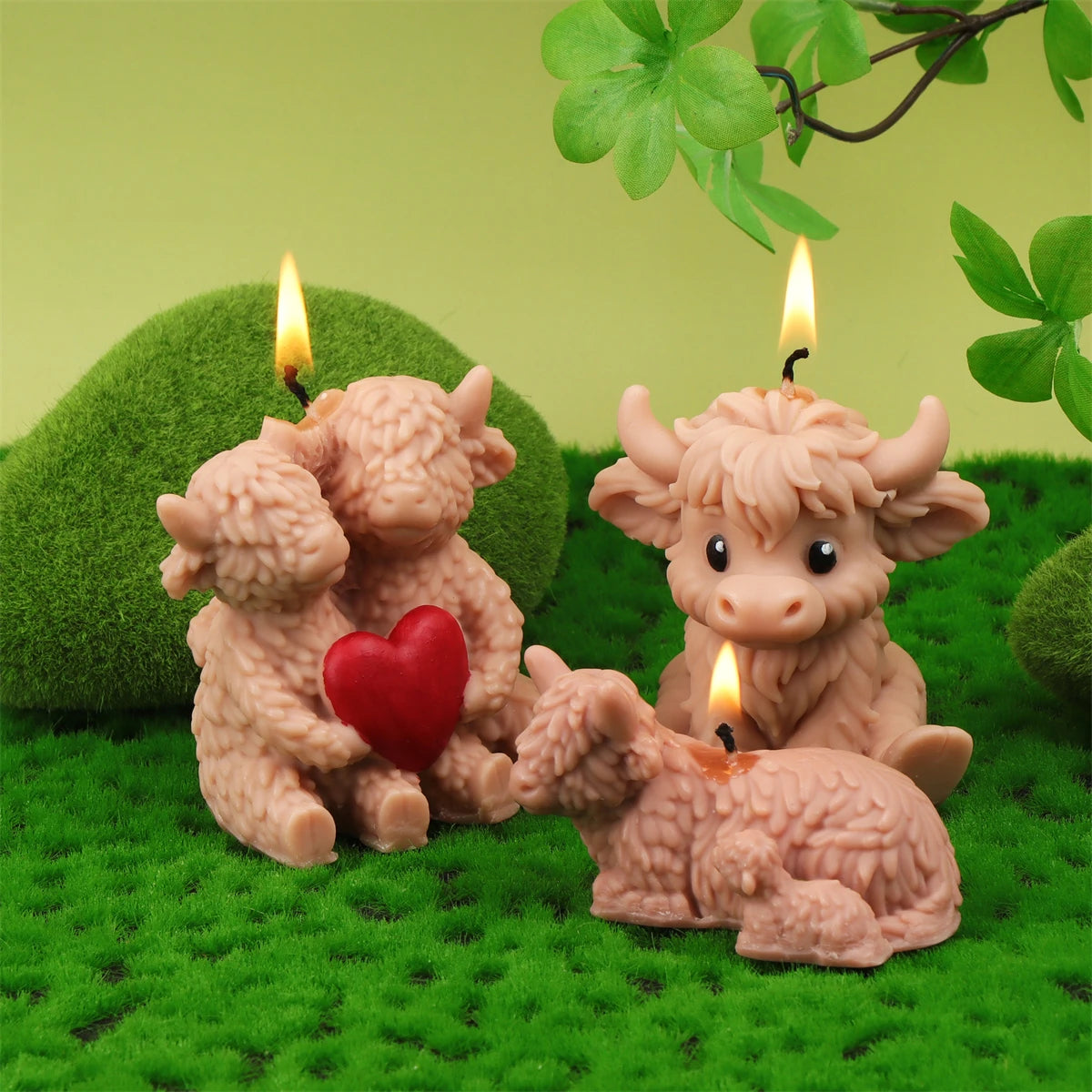 A set of four silicone candle molds shaped like Highland cows, with three lit candles resting on a green grassy surface.