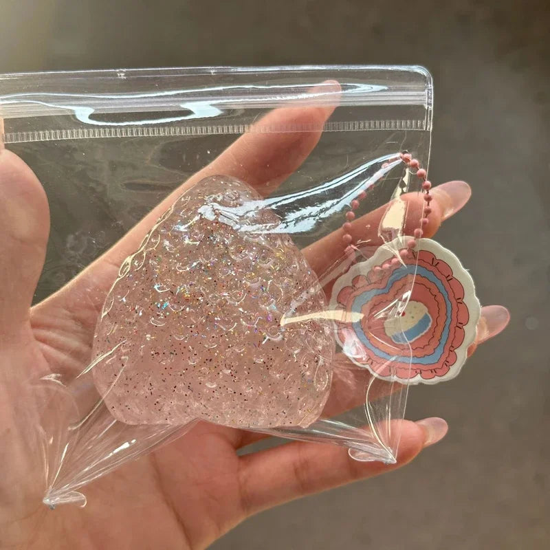 A hand holds a pink glittery squishy fidget toy with a decorative pendant inside a clear plastic bag.