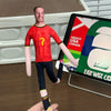 A male figure dressed in a red football jersey with a yellow number, standing in front of a sign displaying flags for Mexico,