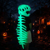 A glowing green dinosaur skeleton mask stands in a dark outdoor setting with autumn foliage and pumpkins.