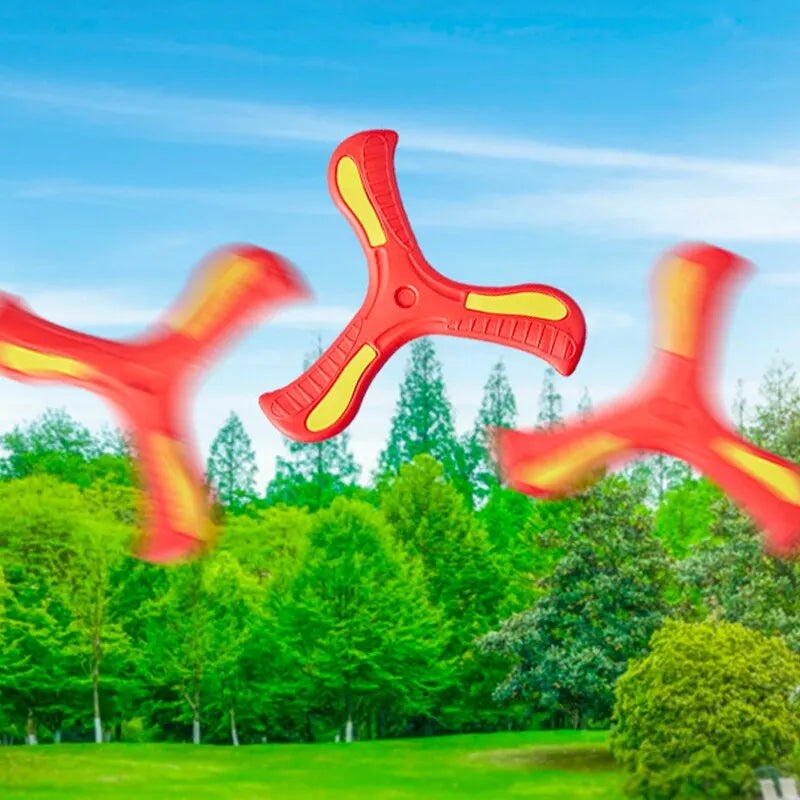 A bright orange soft foam boomerang flying disc is shown against a backdrop of green trees and blue sky.