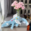 A light blue rainbow dragon plush toy sitting on a gray table next to a vase of pink flowers and wine bottles.