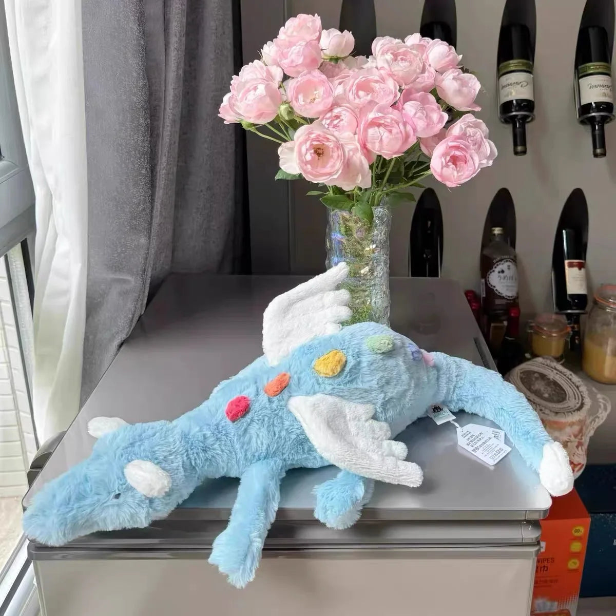 A light blue rainbow dragon plush toy sitting on a gray table next to a vase of pink flowers and wine bottles.