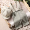 A set of a light sage green knit romper and matching newsboy cap laid on a wooden frame with knitted fabric.