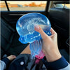 A hand holds a translucent blue jellyfish squish toy with tentacles inside a car interior.