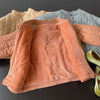 A close-up view of a coral colored padded jacket lying on a dark surface, with other light blue and beige jackets in the back