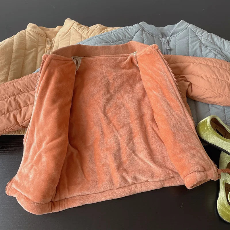 A close-up view of a coral colored padded jacket lying on a dark surface, with other light blue and beige jackets in the back