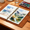 A watercolor sketchbook with lay-flat binding and a palette of paints is displayed on a wooden table.