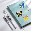 A teal notebook features diamond painting stickers of blue and yellow butterflies, with a silver ring and a pen nearby.