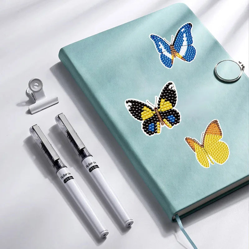 A teal notebook features diamond painting stickers of blue and yellow butterflies, with a silver ring and a pen nearby.
