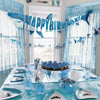 A blue and white geometric shark happy birthday banner and hanging garland set decorating a table.