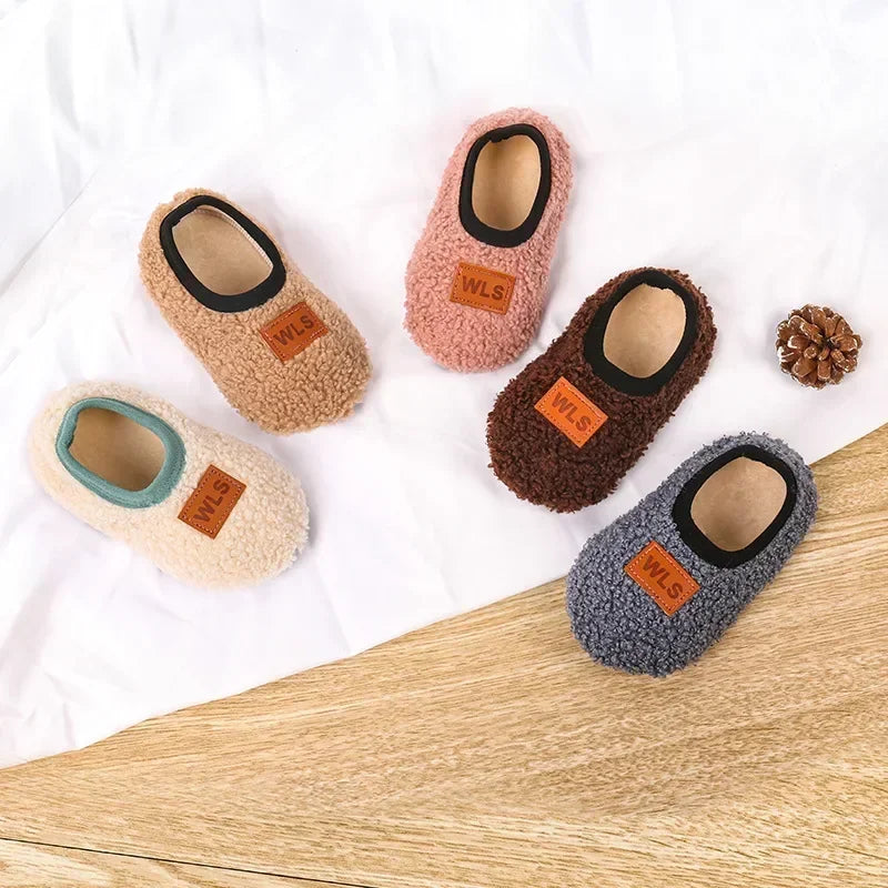 A collection of five plush baby slippers in various colors including pink, brown, beige, and gray, displayed on a white surfa