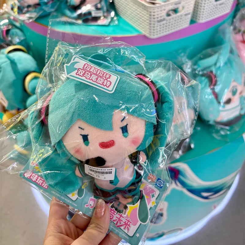 A teal Hatsune Miku plush keychain with pink accents is displayed on a tiered stand.