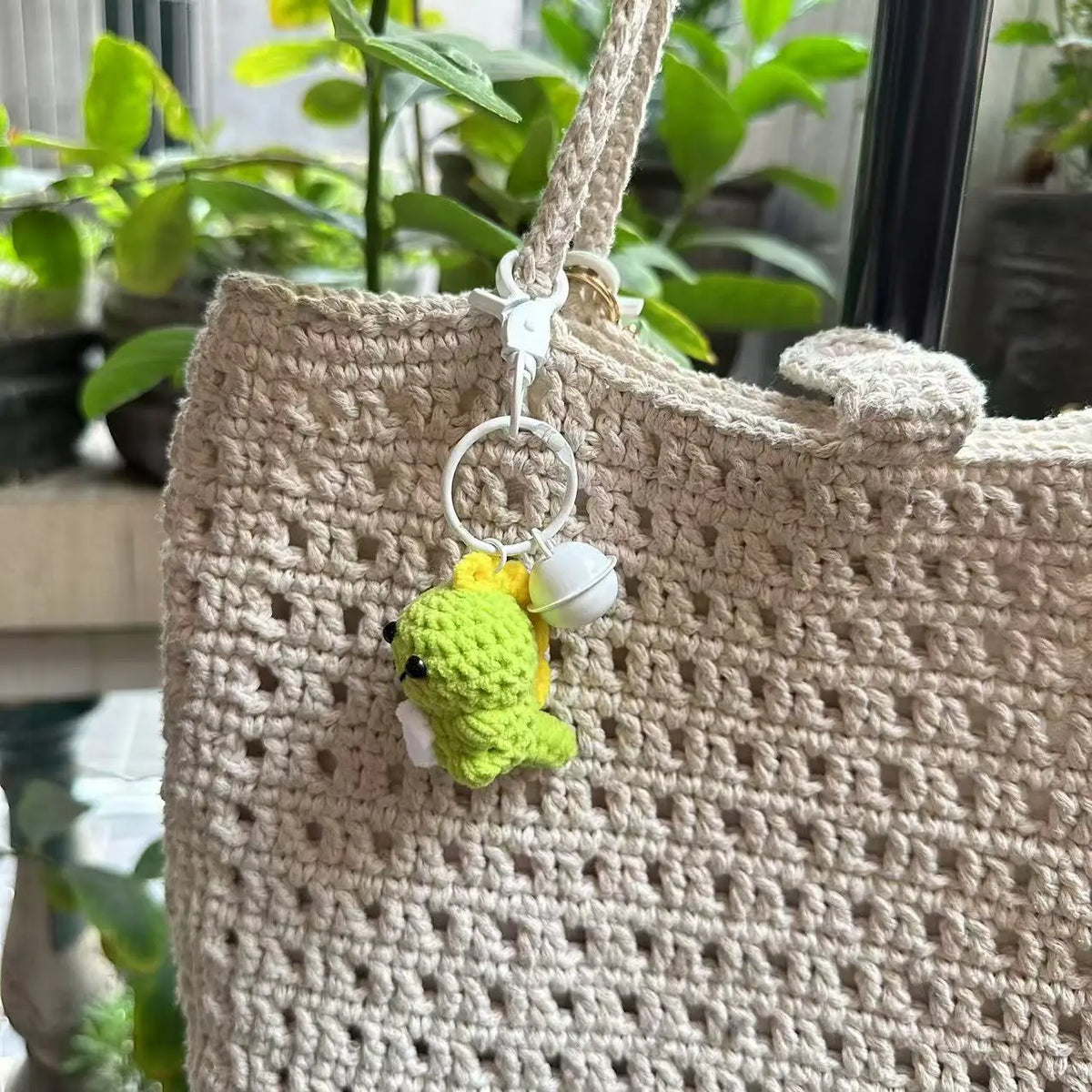 A handmade crochet dinosaur keychain hanging from a light beige woven bag.