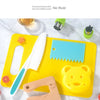 A yellow cutting board holds a white plastic knife, a blue plastic knife, and a yellow plastic bear shape.