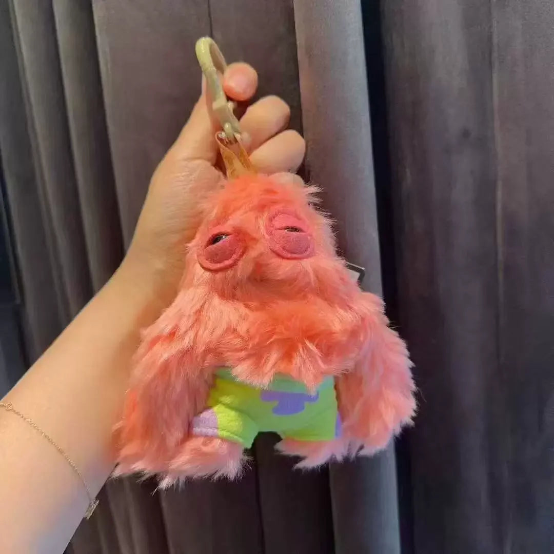 A fluffy orange monster plush toy with large eyes and a green outfit being held by a hand.