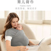 A woman is holding a baby in an ergonomic sling carrier while reading a book indoors.
