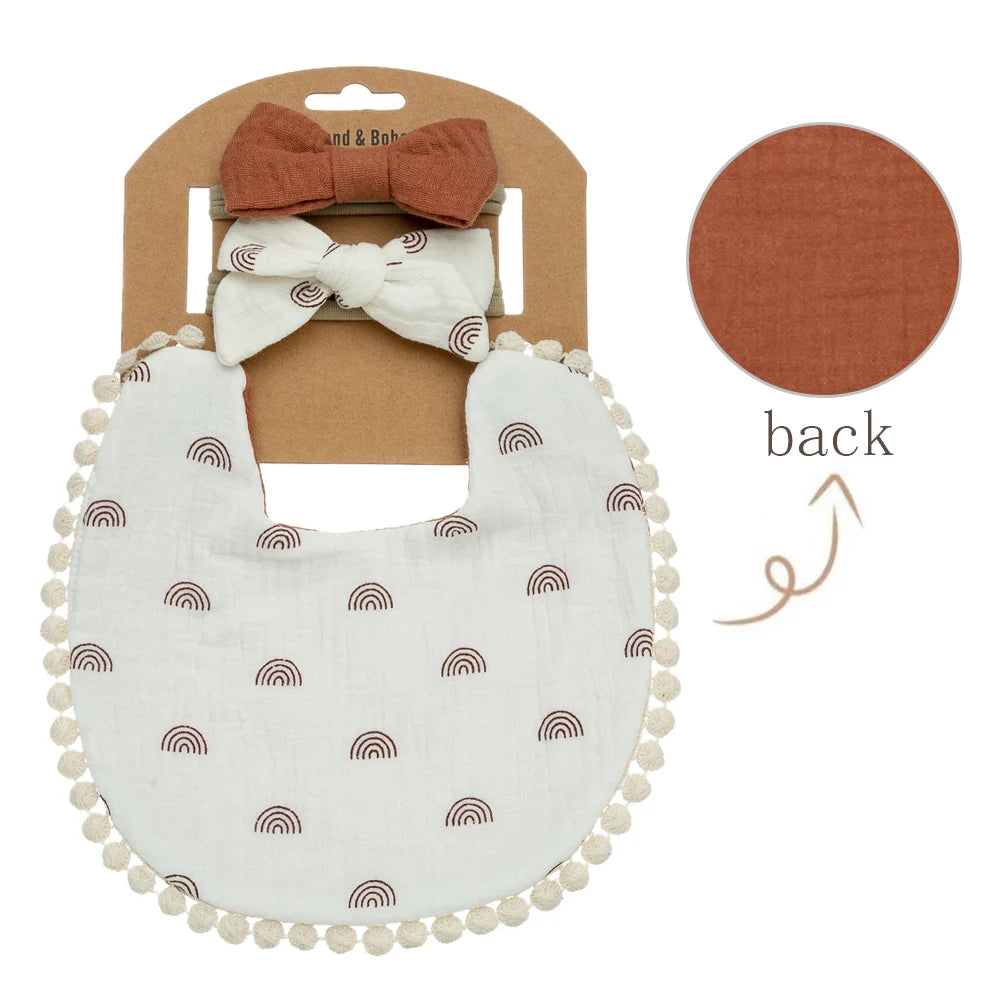 A white fabric bib with a rainbow pattern and a matching brown bow headband shown next to its packaging.