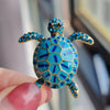 A turquoise and gold rhinestone turtle brooch pin is held by a person's fingers.