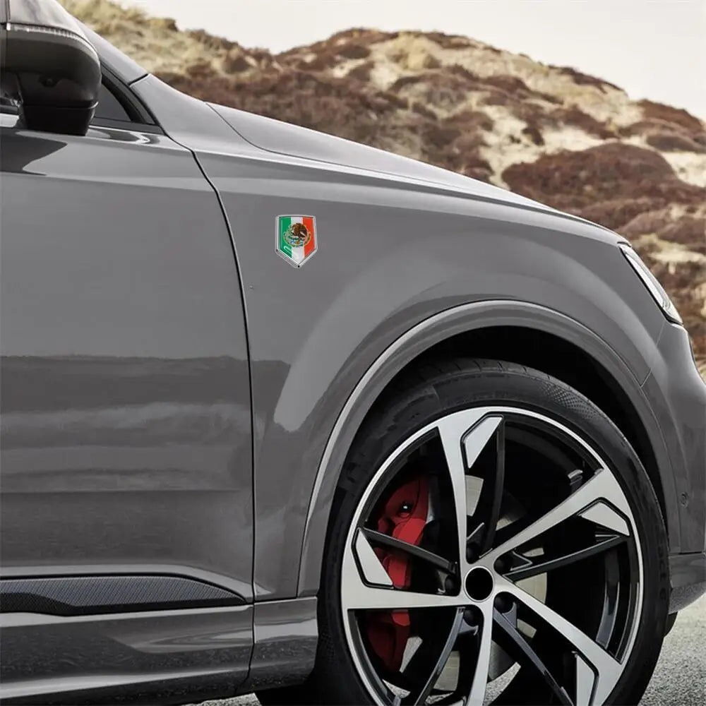 A close-up of a gray SUV featuring a 3D chrome Mexican flag emblem decal on the hood and a black alloy wheel.