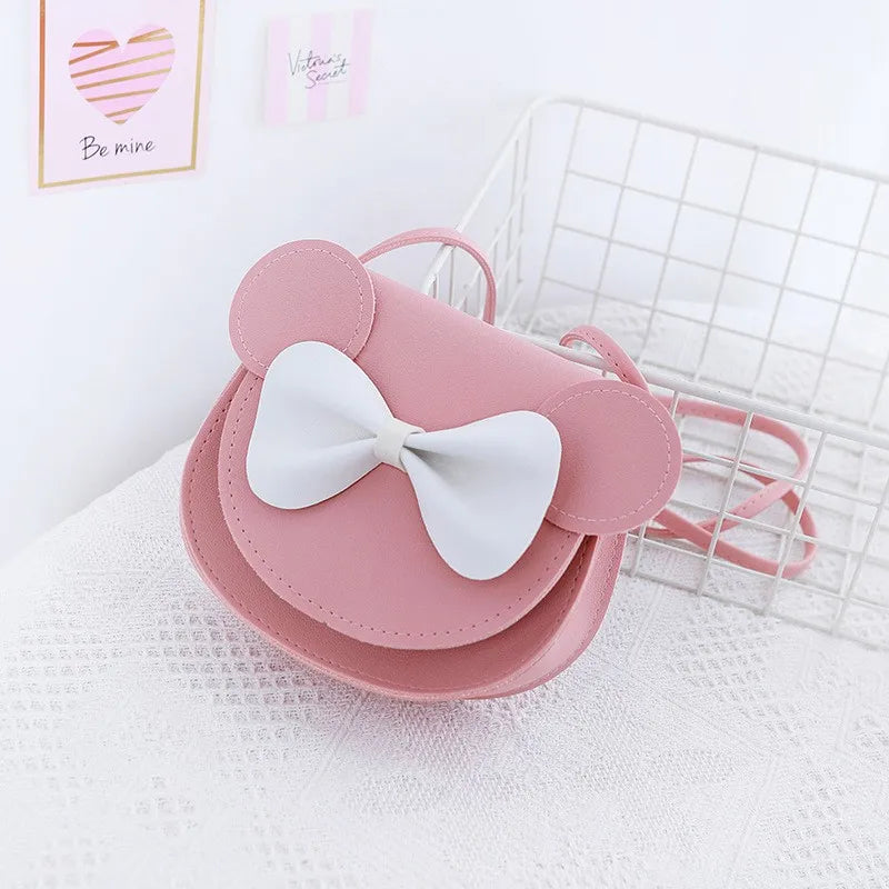 A pink kids' hard shell crossbody bag featuring mouse ears and a white bow, resting on a white textured surface.