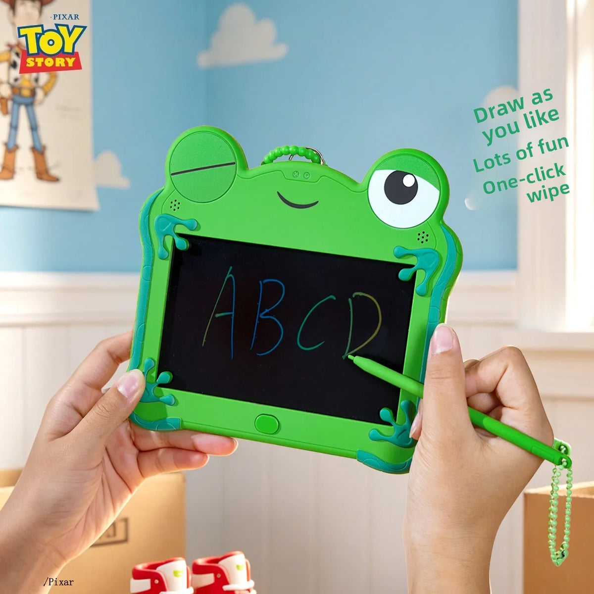 A child holds a green frog LCD drawing tablet with a black screen and a pencil, demonstrating an educational toy.