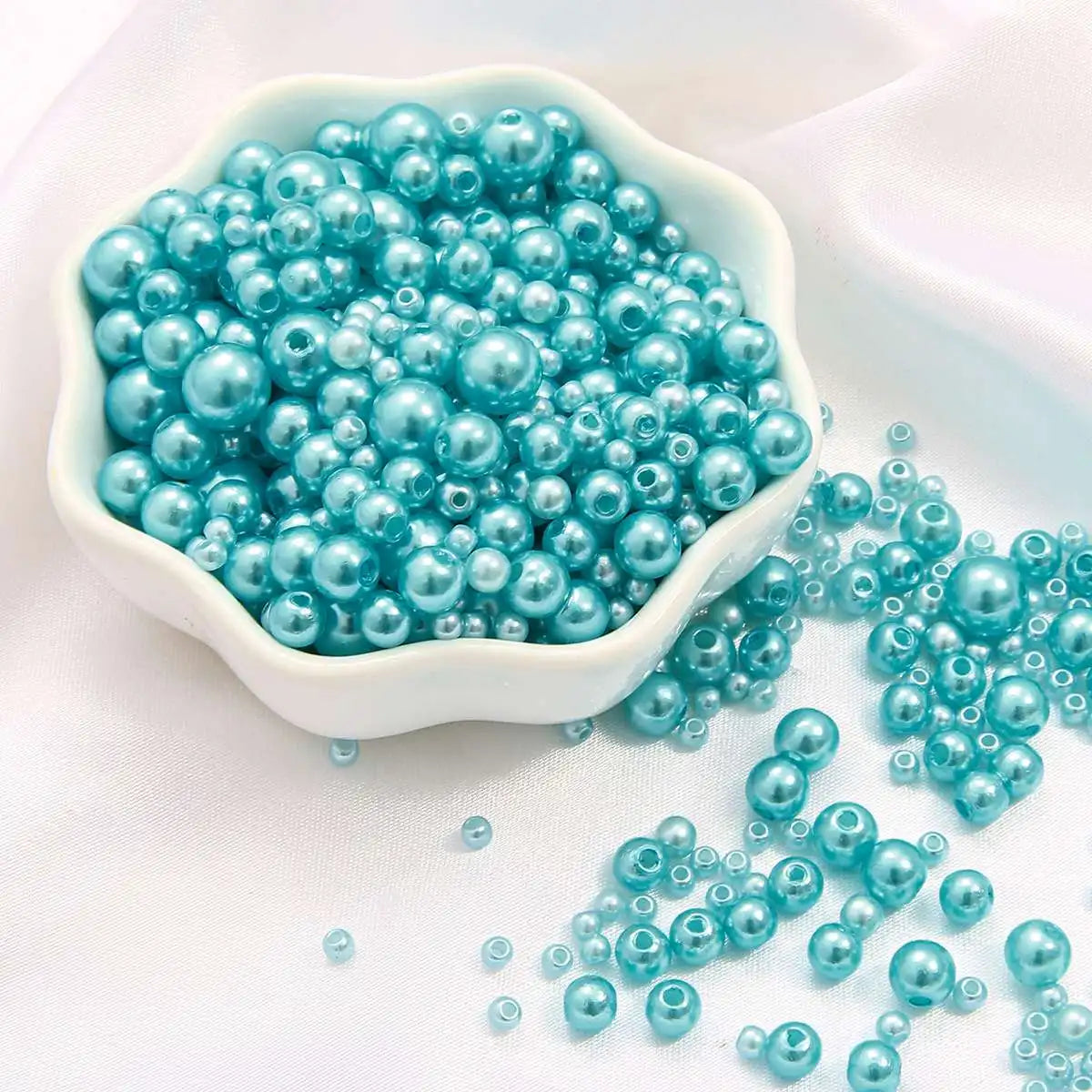 A white bowl filled with assorted teal blue pearl beads, some spilled onto a white surface.