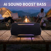 A black portable Bluetooth speaker with purple accent lighting is placed on a wooden table outdoors near a campfire.