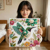 A woman holds a finished diamond painting of a vibrant green and black bird adorned with pearls and diamonds.