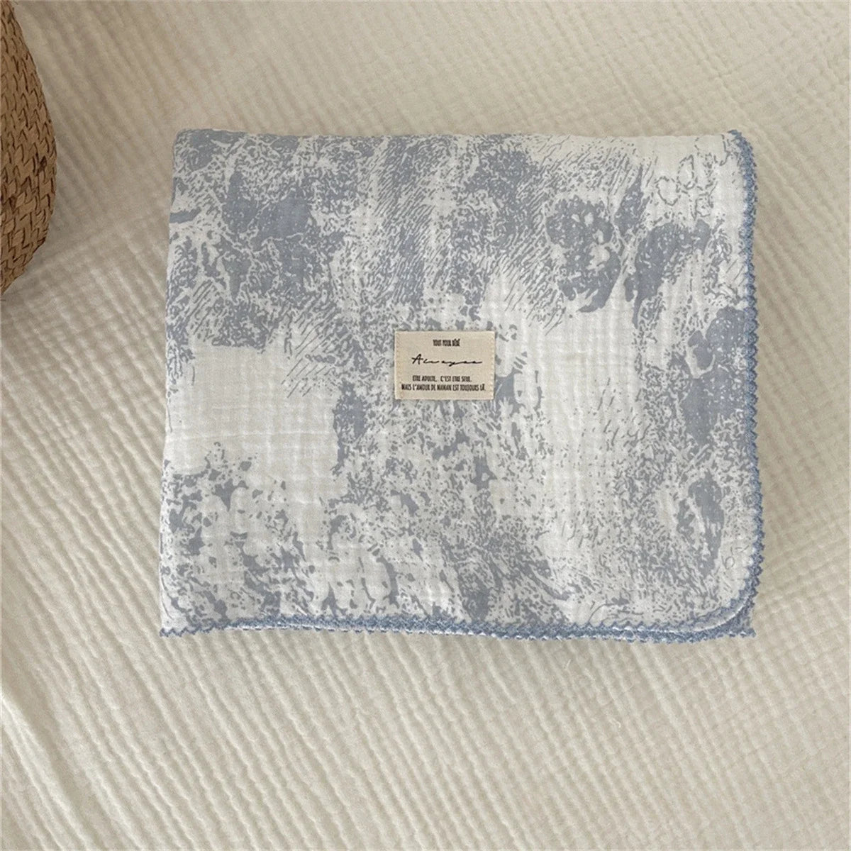 A light blue muslin swaddle blanket with a subtle floral print is displayed on a cream-colored fabric.