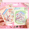 A hand holds an open 21-piece miniature cottage 3D sticker book featuring colorful illustrations for journaling and crafts.