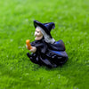 A black witch figurine with a white hat and flowing cloak is lying on green grass.