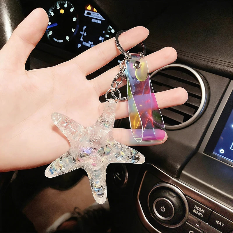 A hand holds a clear starfish charm attached to a colorful, iridescent pendant keychain.