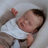 A newborn baby doll with rooted hair is sleeping in a white, textured cradle.