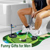 A man is sitting on a toilet while practicing indoor putting with a mat, putter, and balls.