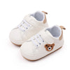 A pair of cream-colored baby sneakers with white laces and a brown bear appliqué.