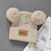 A beige knit baby beanie hat with fluffy pom-poms on the ears and a brown patch on the front.