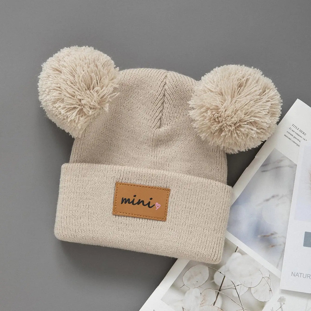 A beige knit baby beanie hat with fluffy pom-poms on the ears and a brown patch on the front.