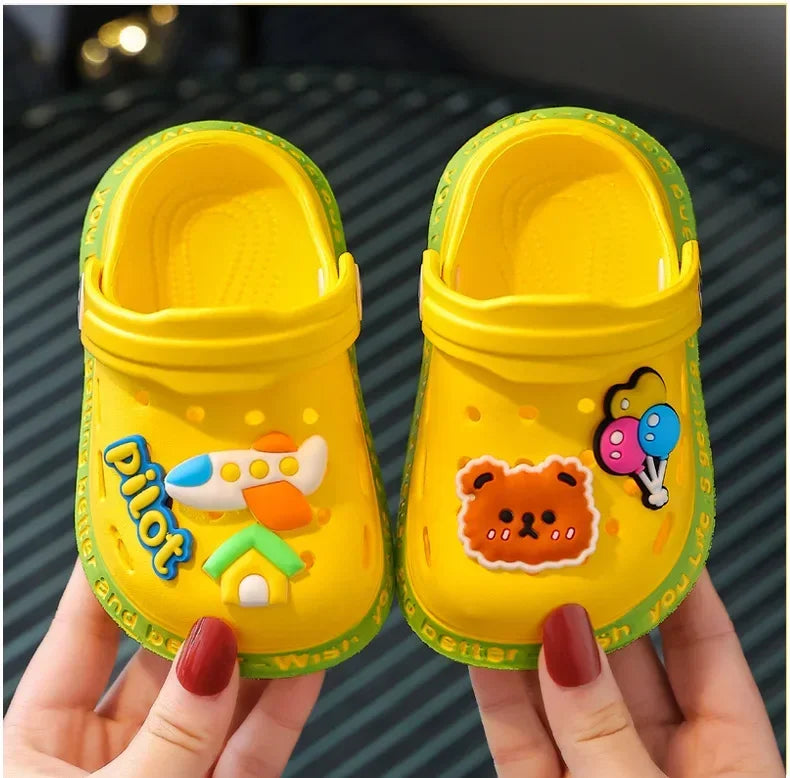 A pair of bright yellow children's clogs with orange and green accents are held by a hand.