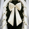 Oversized creamy white satin bow decoration hanging in a white archway decorated with white flowers.