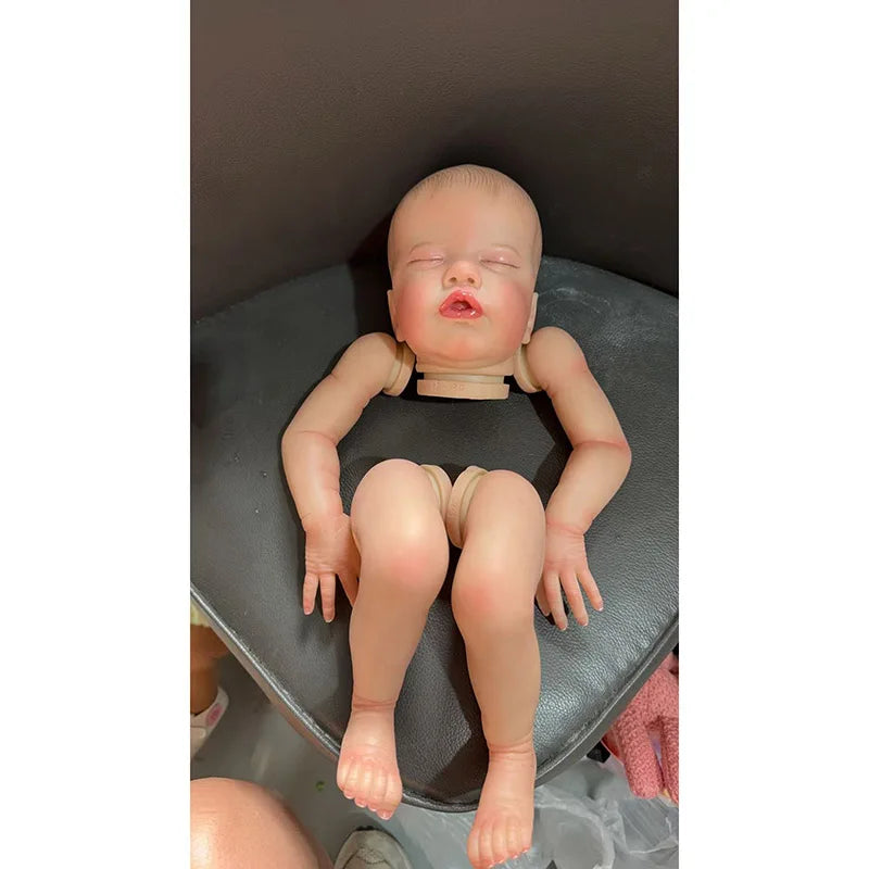 A realistic reborn baby doll kit with painted details and a cloth body is shown lying on a dark surface.